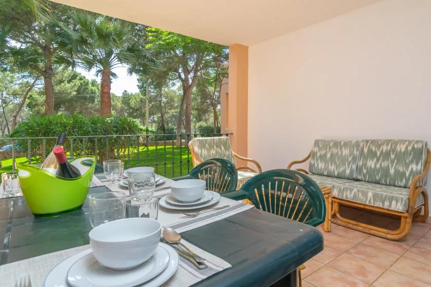 Una zona cubierta para comer al aire libre, con la mesa puesta y asientos acolchados, da a un jardín frondoso.