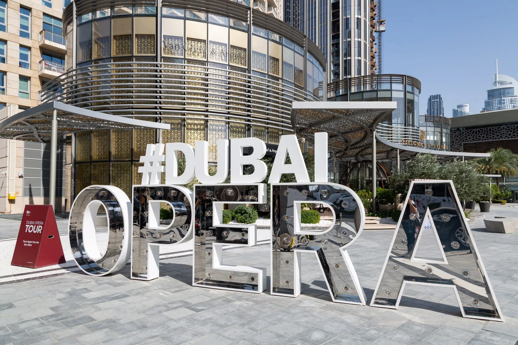 Large “Dubai Opera” letters stand in a paved plaza outside a modern building.