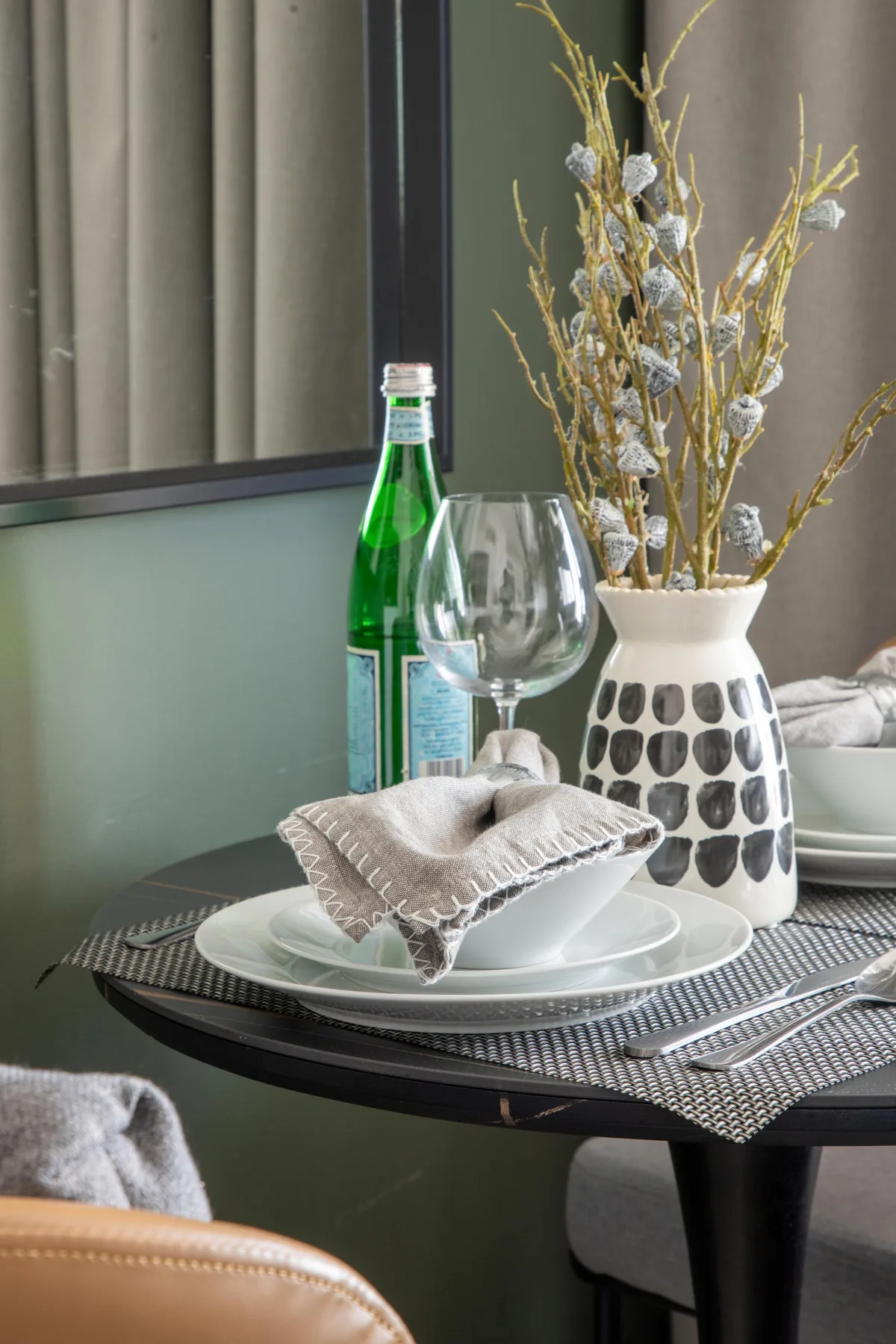 A dining table is set with plates, a glass, and a vase of dried branches.