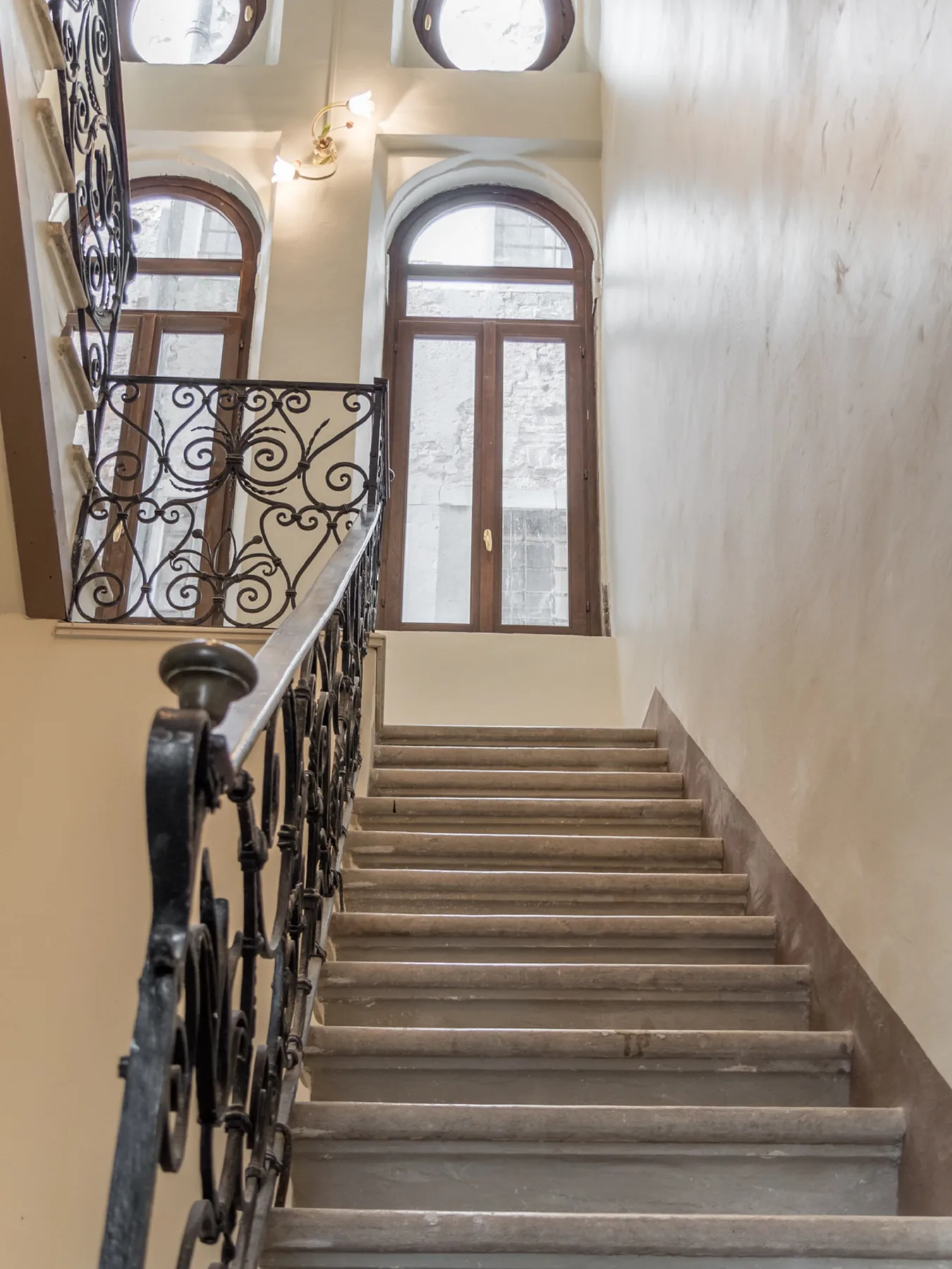 Una escalera interior con barandales decorativos oscuros y ventanas altas en arco.