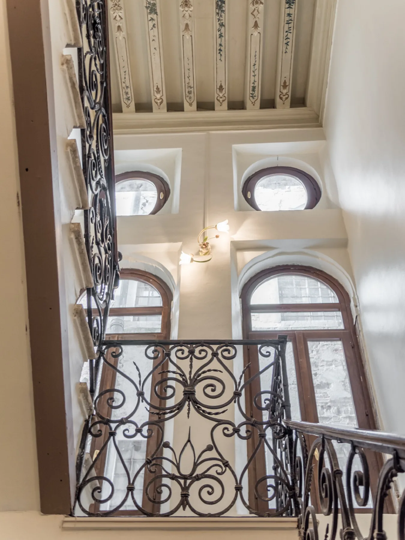 Una escalera interior con barandales ornamentados, ventanas altas en arco y techo decorado.