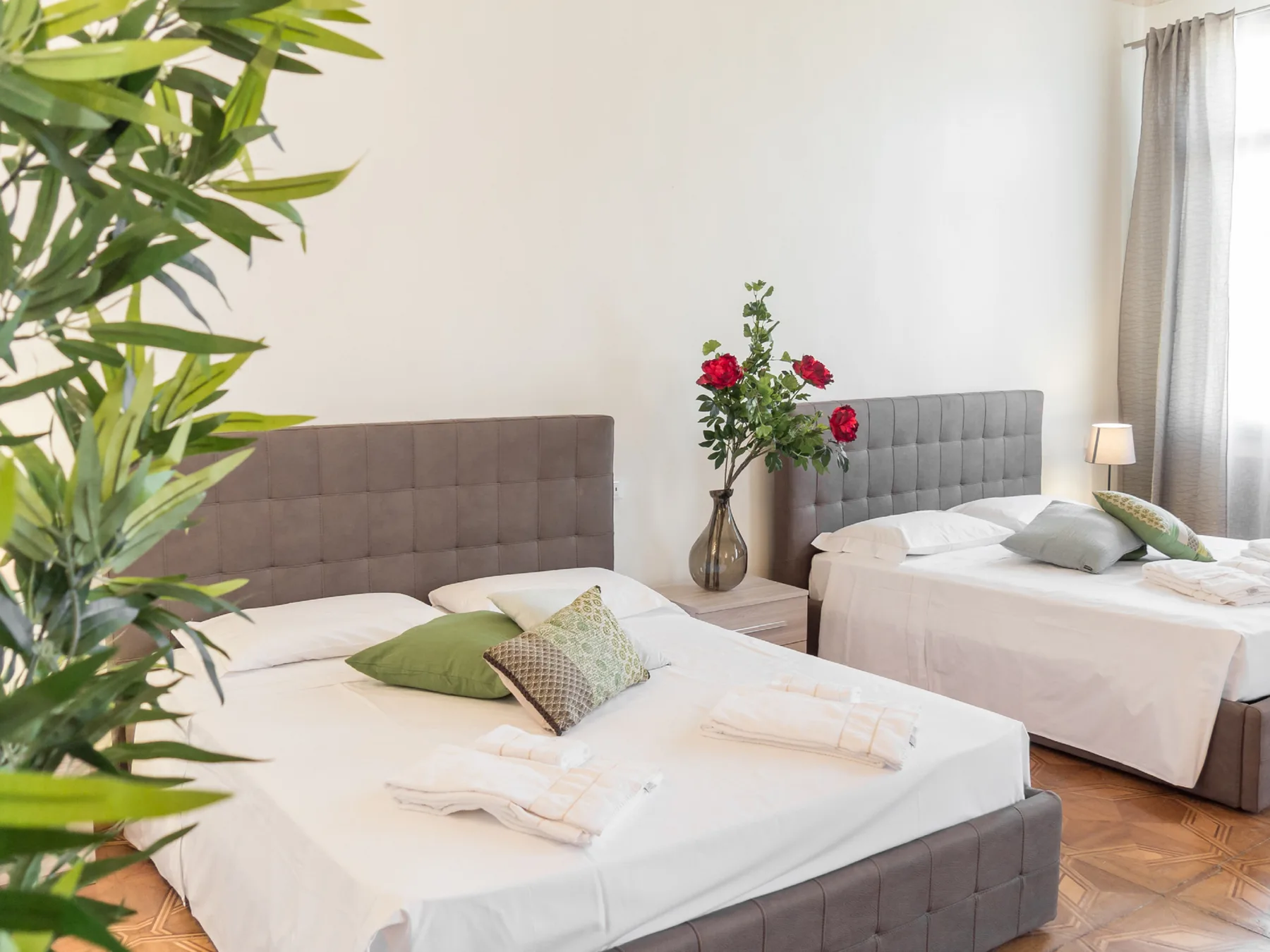 Un dormitorio luminoso con dos camas, ropa de cama neutra y un jarrón con flores.