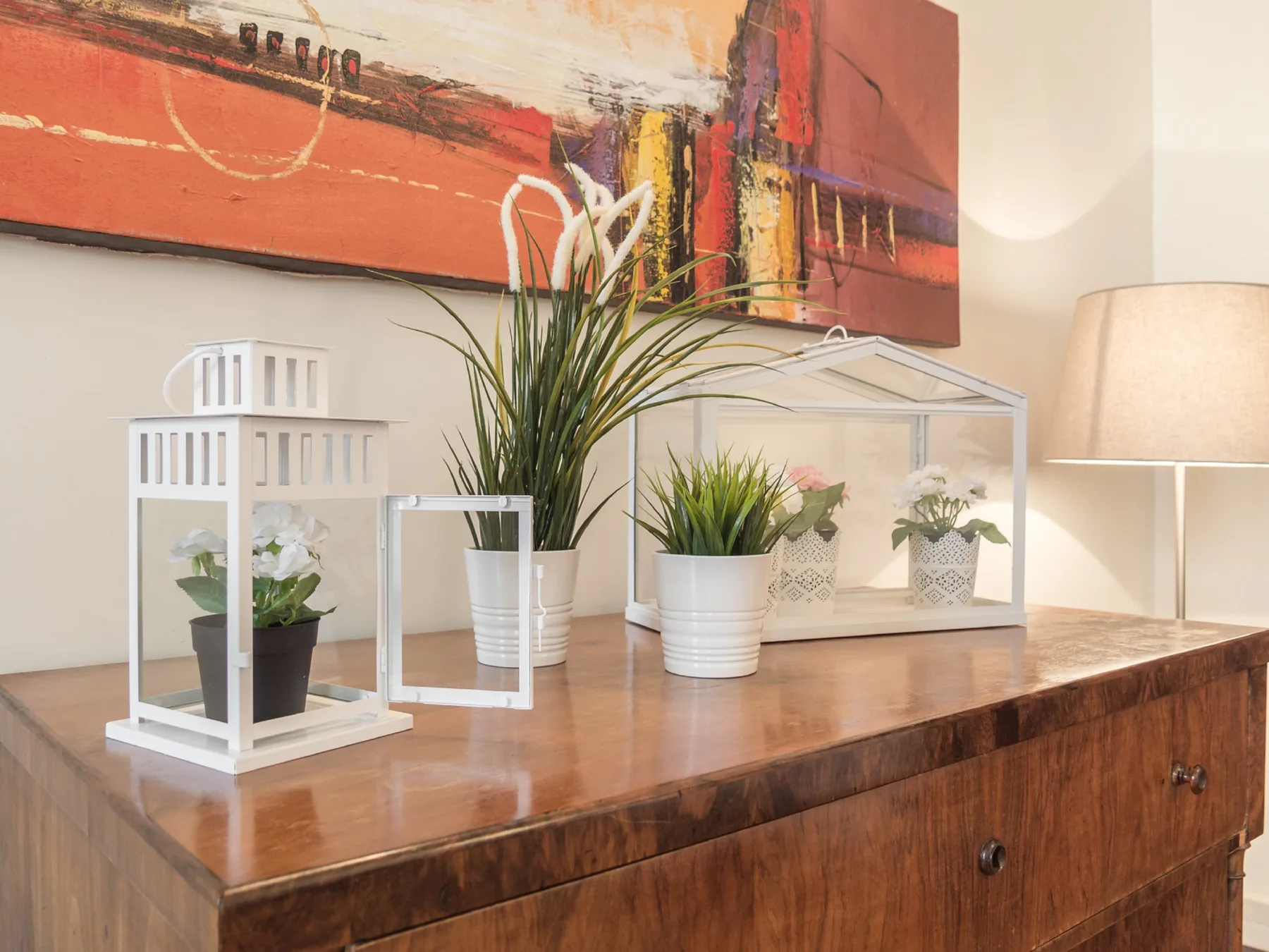 Una consola de madera exhibe faroles decorativos, plantas en macetas y una pintura abstracta.