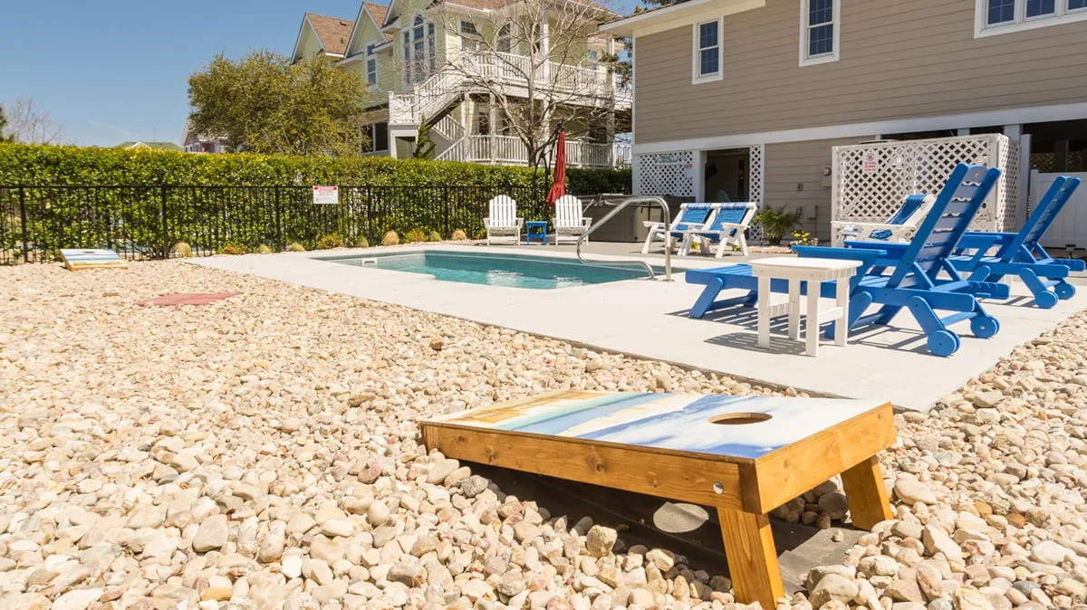 An outdoor pool area has blue lounge chairs and a sandy play area.