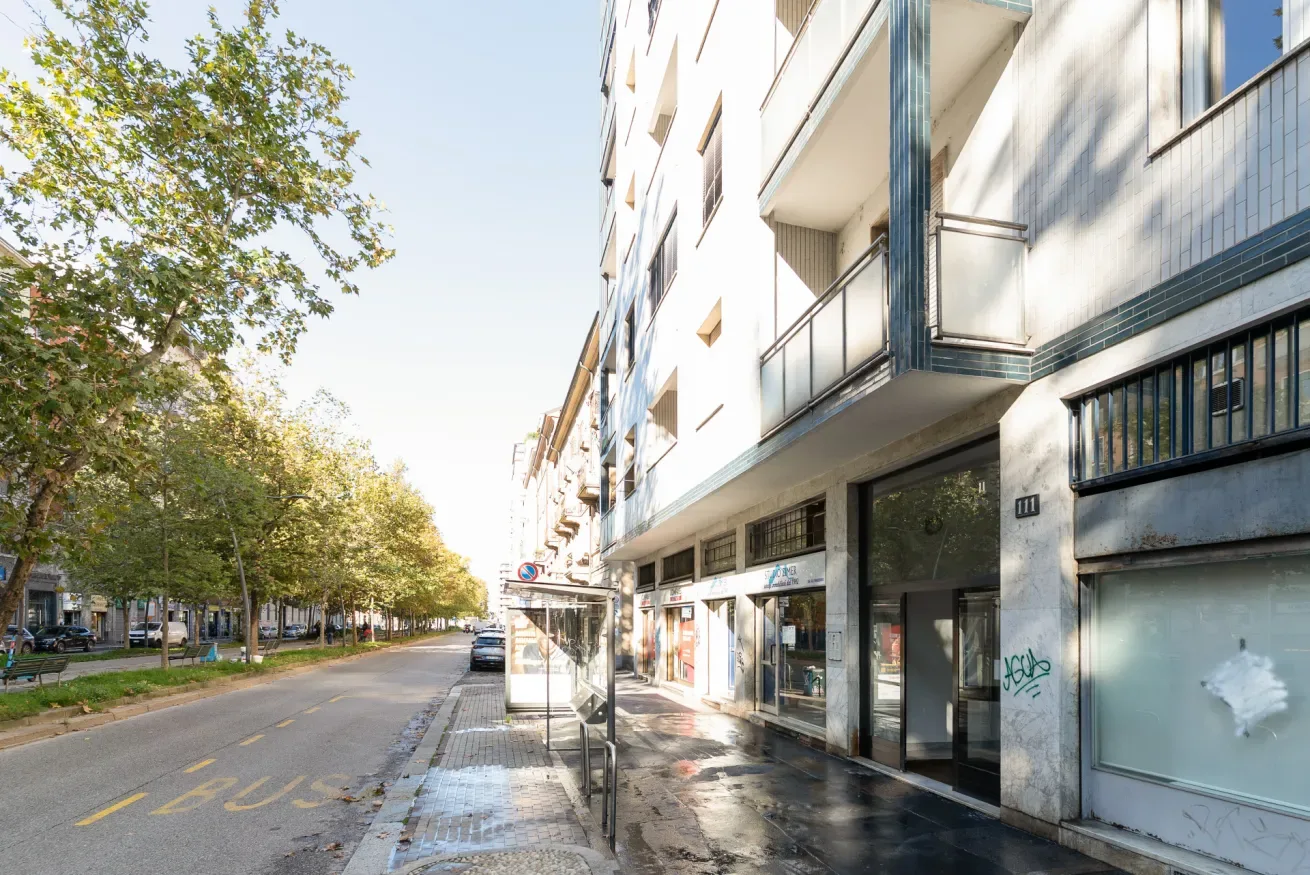 A city street runs alongside a multistory building with balconies and storefronts.