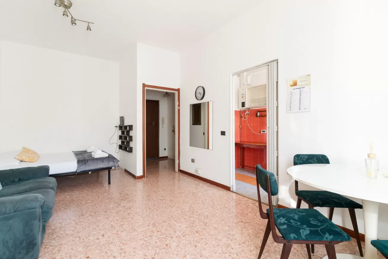 A bright room with a bed, dining table, and open doorway to a red kitchen.