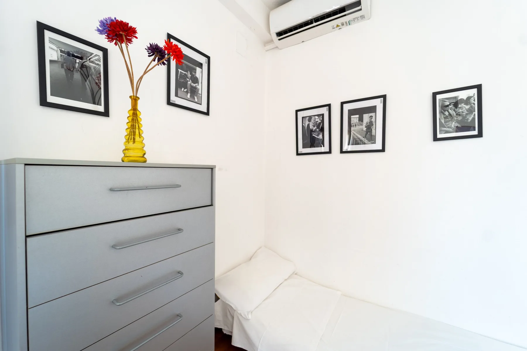 Una esquina luminosa de dormitorio con cama, cómoda, cuadros enmarcados y flores.