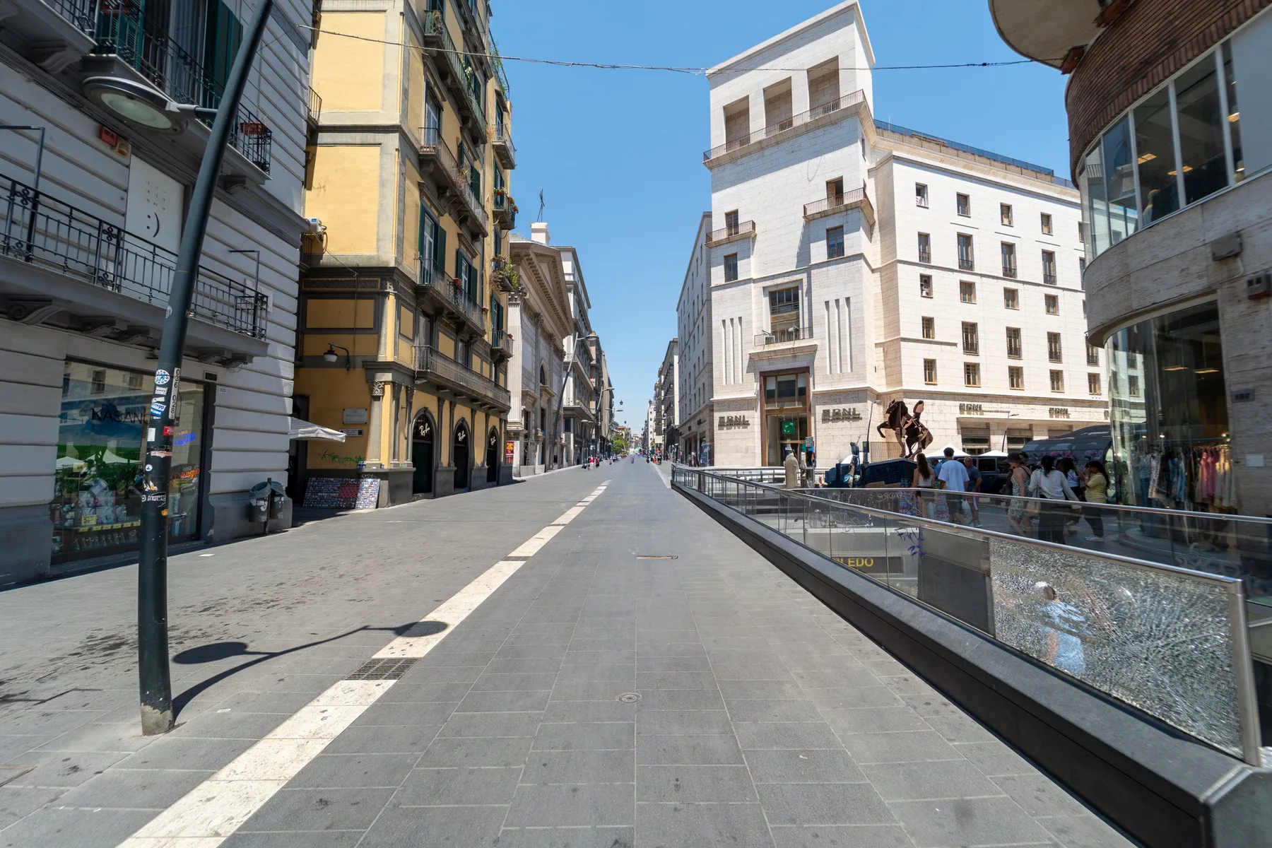 Una calle peatonal pavimentada discurre entre edificios de varias plantas bajo un cielo azul despejado.