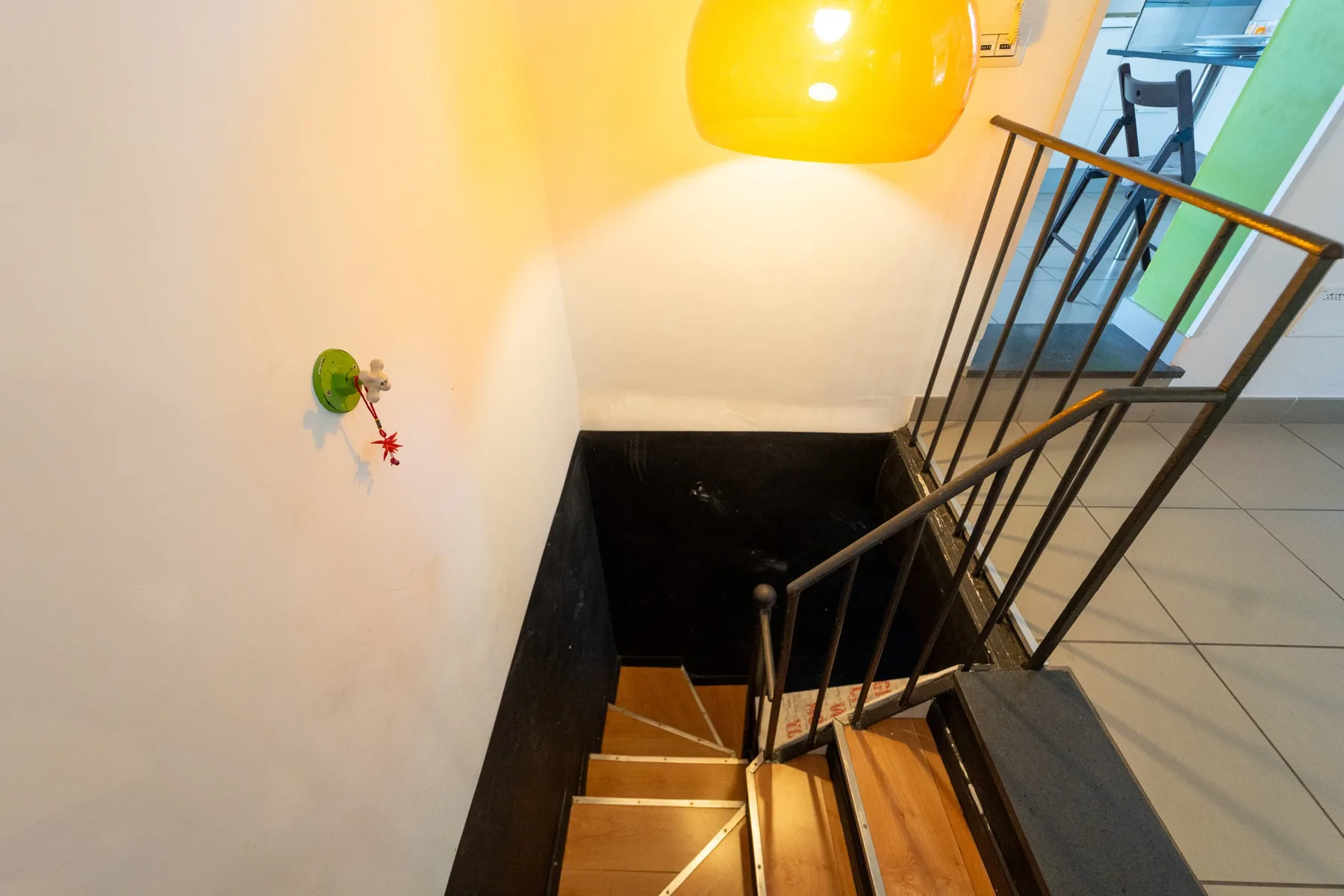 Una escalera interior desciende junto a una barandilla metálica negra bajo una lámpara amarilla.