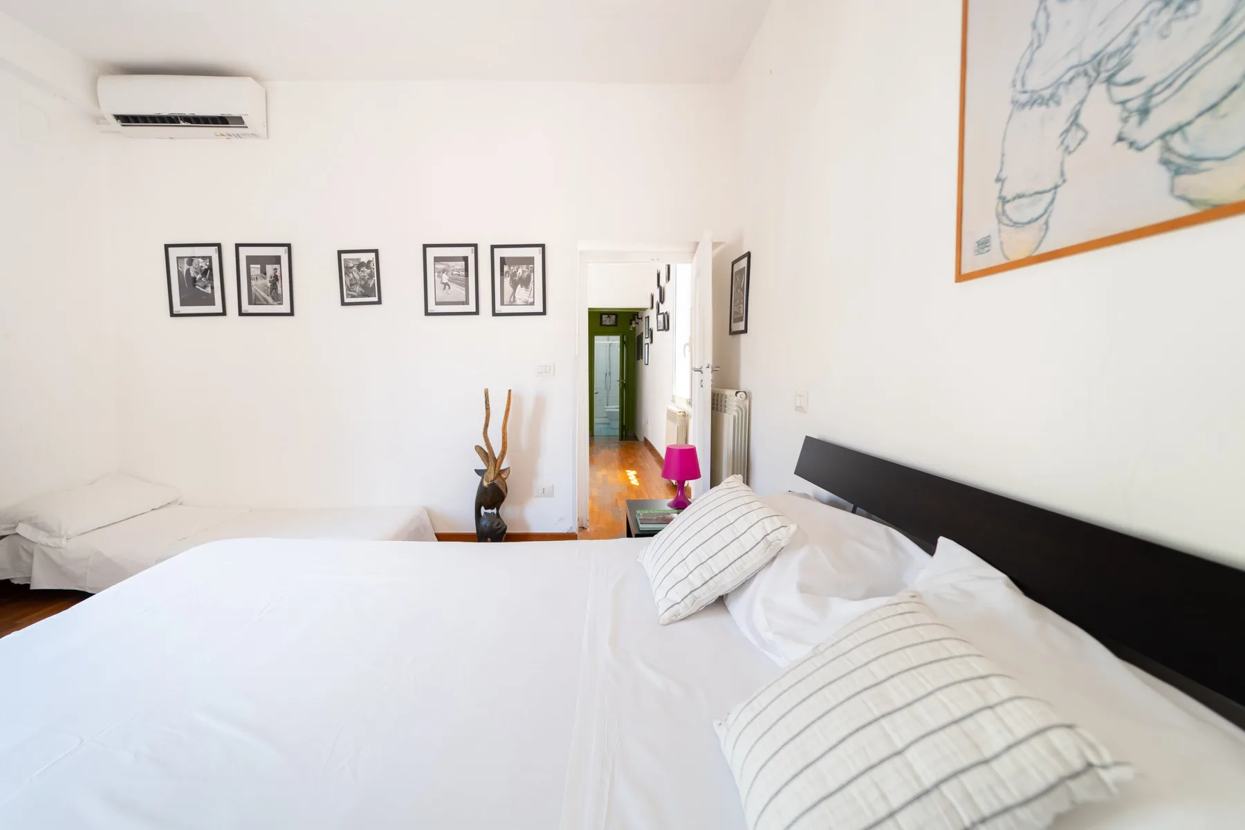 Un dormitorio luminoso con ropa de cama blanca, cuadros enmarcados y una puerta hacia el pasillo.
