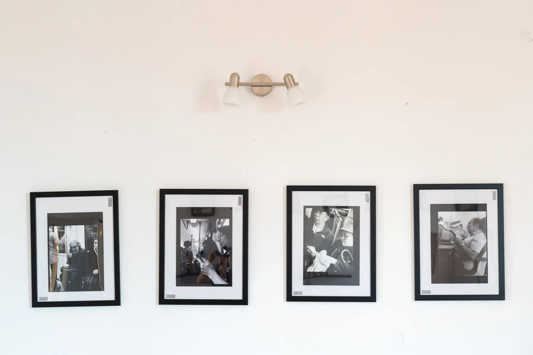 Una pared blanca muestra una fila de fotografías enmarcadas en blanco y negro bajo una pequeña lámpara.