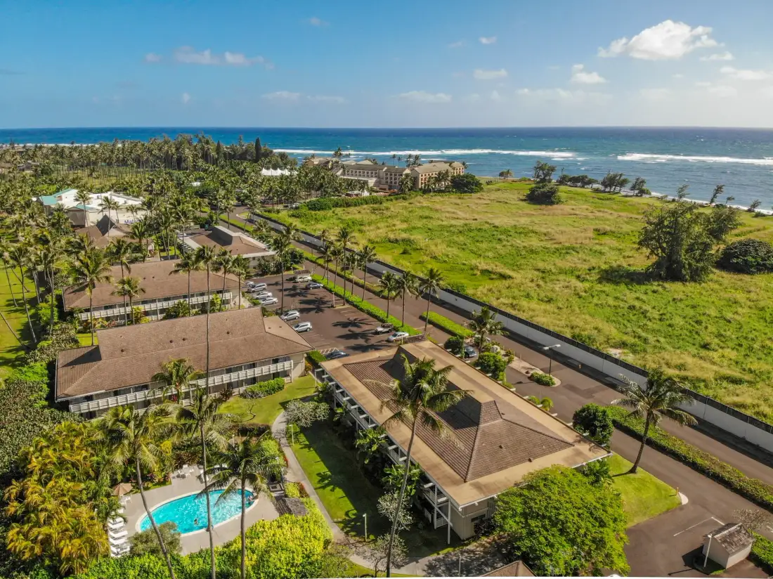 Aerial view of a coastal resort with low buildings, a swimming pool, and ocean beyond.