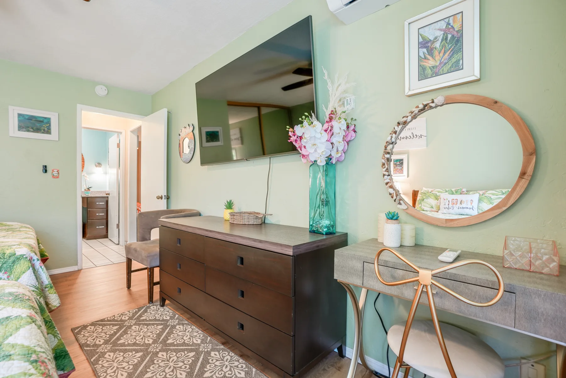 A bedroom with a dark dresser, a wall-mounted television, and a desk beneath a round mirror.