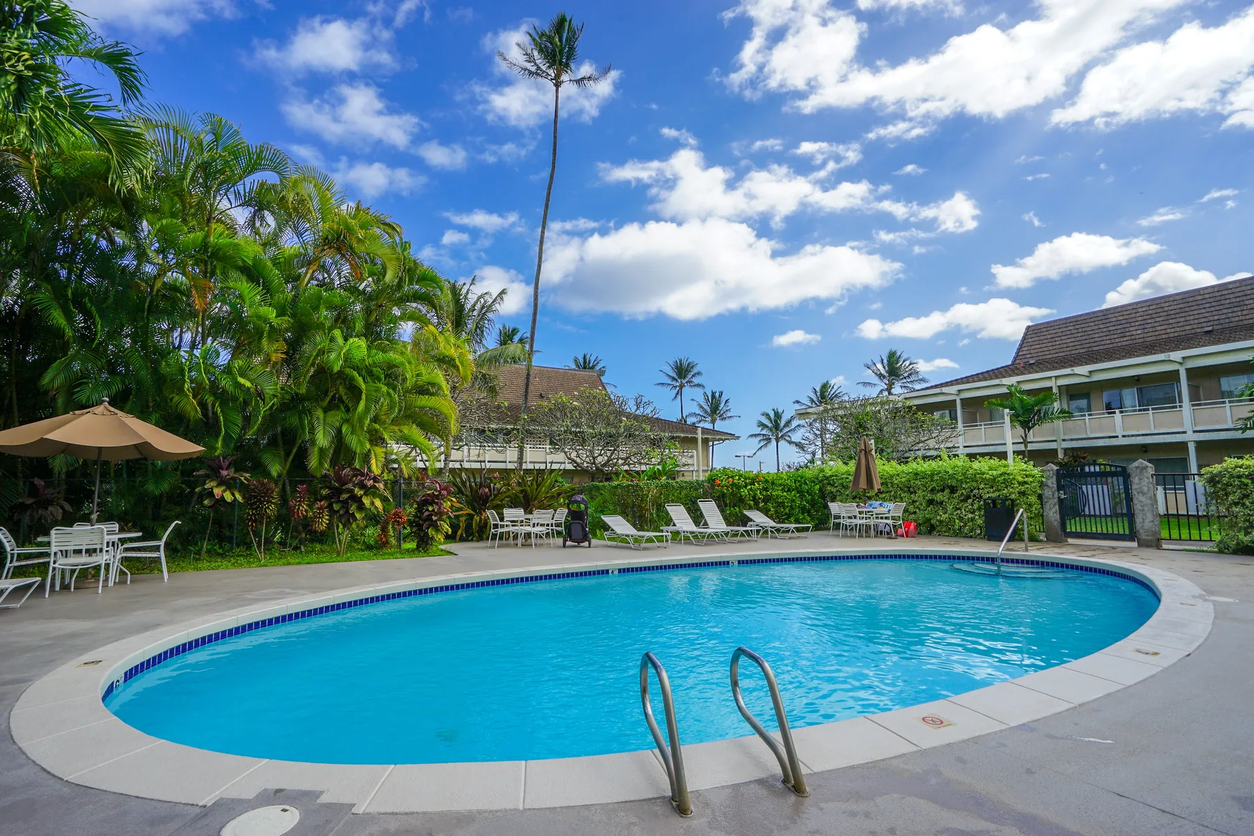 An outdoor swimming pool sits among tropical landscaping and low-rise buildings.