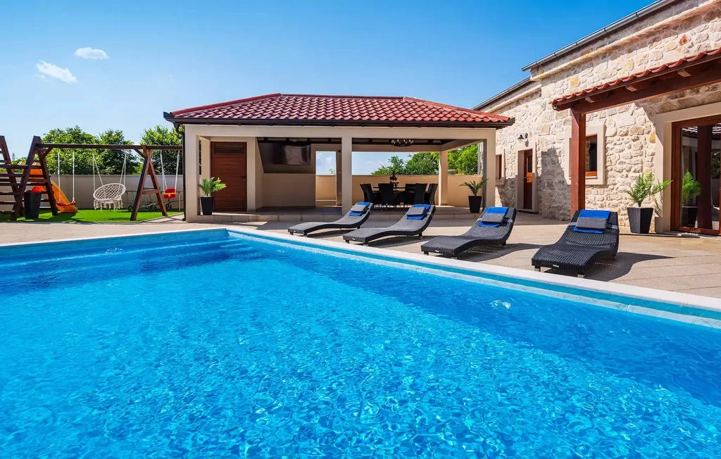 A swimming pool beside a stone house, with lounge chairs and a covered patio.