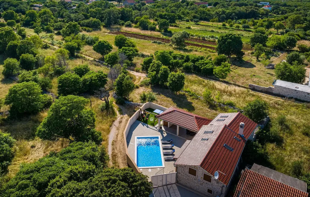 An aerial view shows a house with a blue swimming pool amid green countryside.