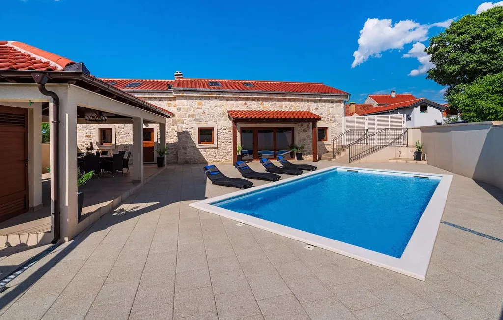 A swimming pool sits beside a stone house and a covered outdoor seating area.