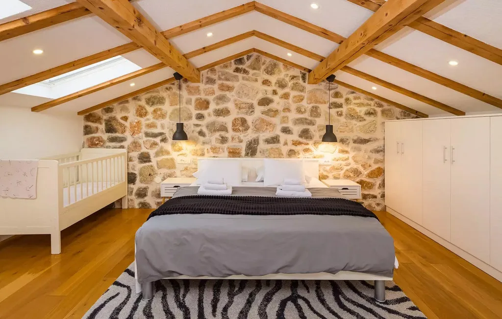 A bedroom with a bed beneath exposed wooden roof beams and a stone wall.