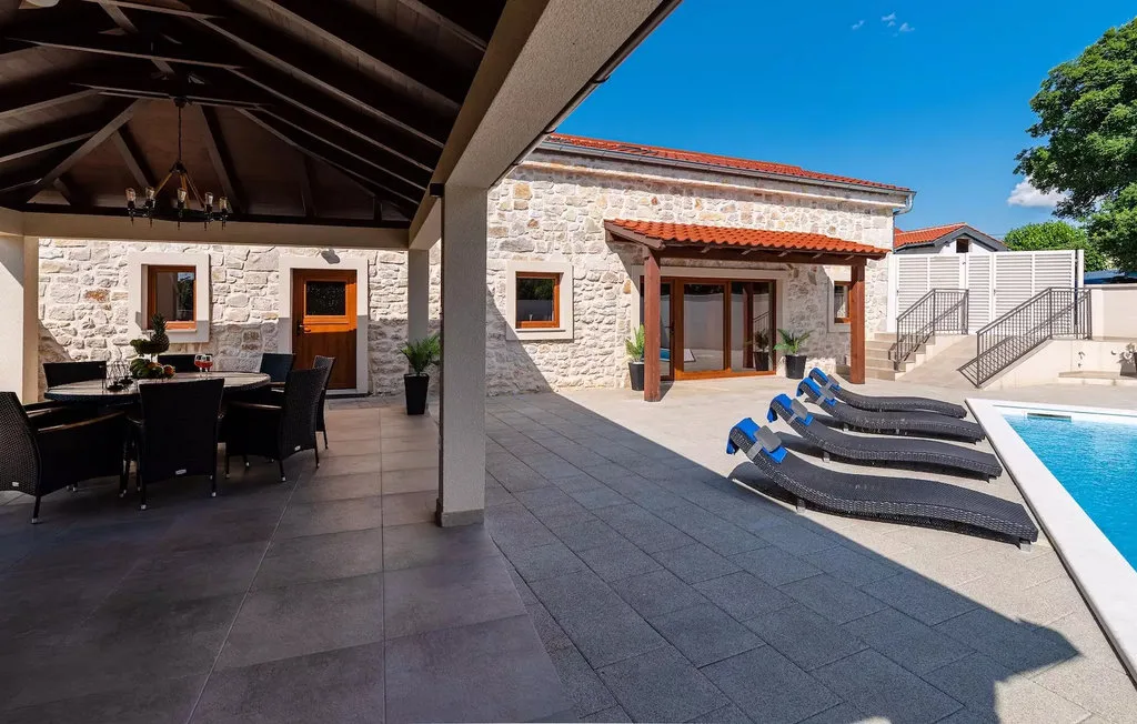 A stone patio with shaded dining seating and lounge chairs beside a swimming pool.
