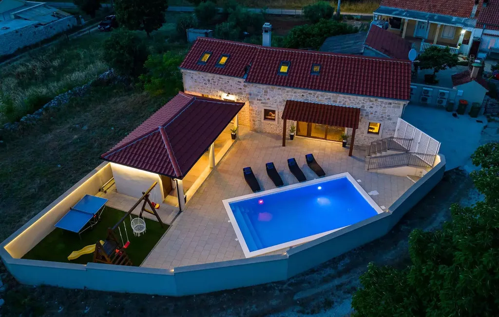 An aerial view of a house, outdoor seating area, and illuminated swimming pool.