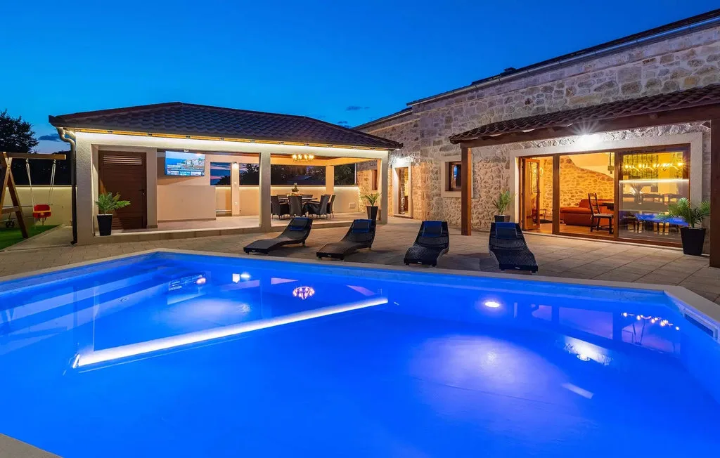 A blue-lit outdoor swimming pool sits beside a stone house at dusk.