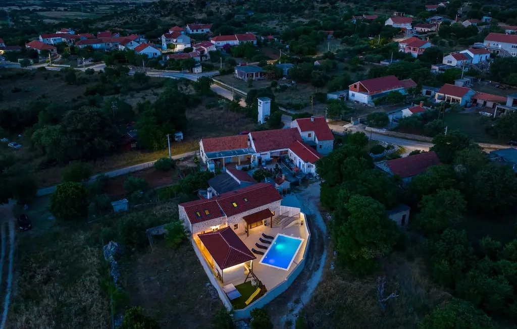 An aerial view shows a lit house and swimming pool among neighboring homes.