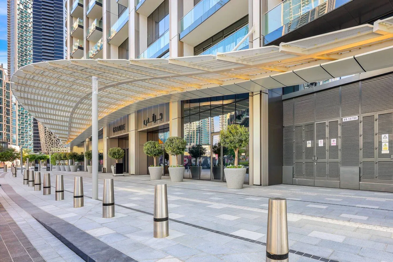 Entrada de un edificio pavimentada bajo una amplia cubierta, con escaparates de vidrio y bolardos metálicos.