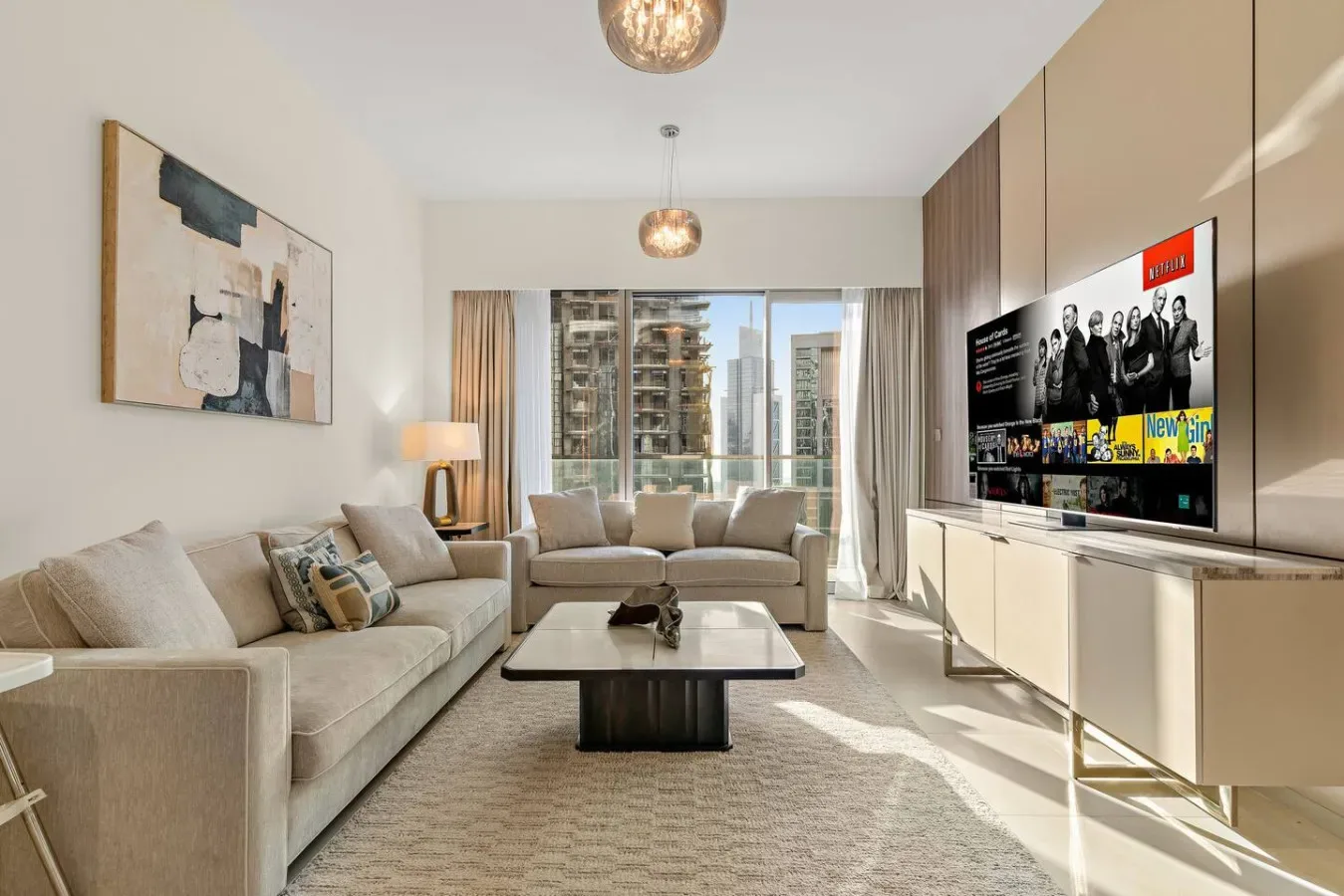 Una sala de estar con sofá, mesa de centro y televisor junto a grandes ventanas.
