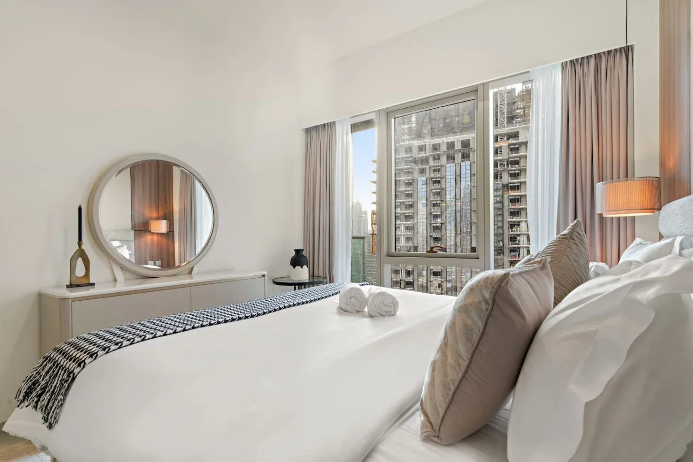 Un dormitorio luminoso con una cama bien tendida y grandes ventanas con vistas a edificios cercanos.