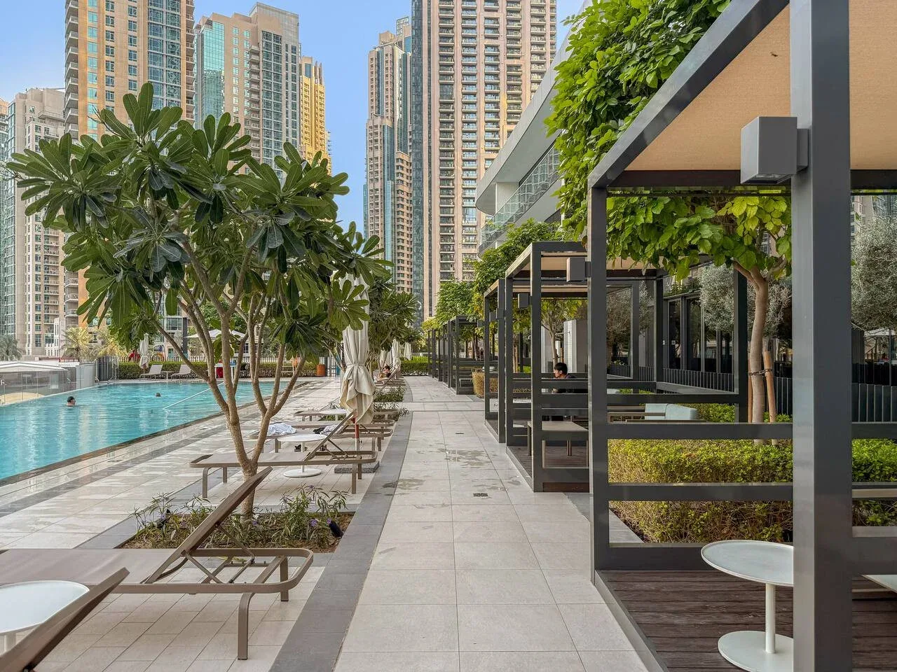 Una terraza junto a la piscina tiene asientos, pérgolas con vegetación y edificios altos al fondo.