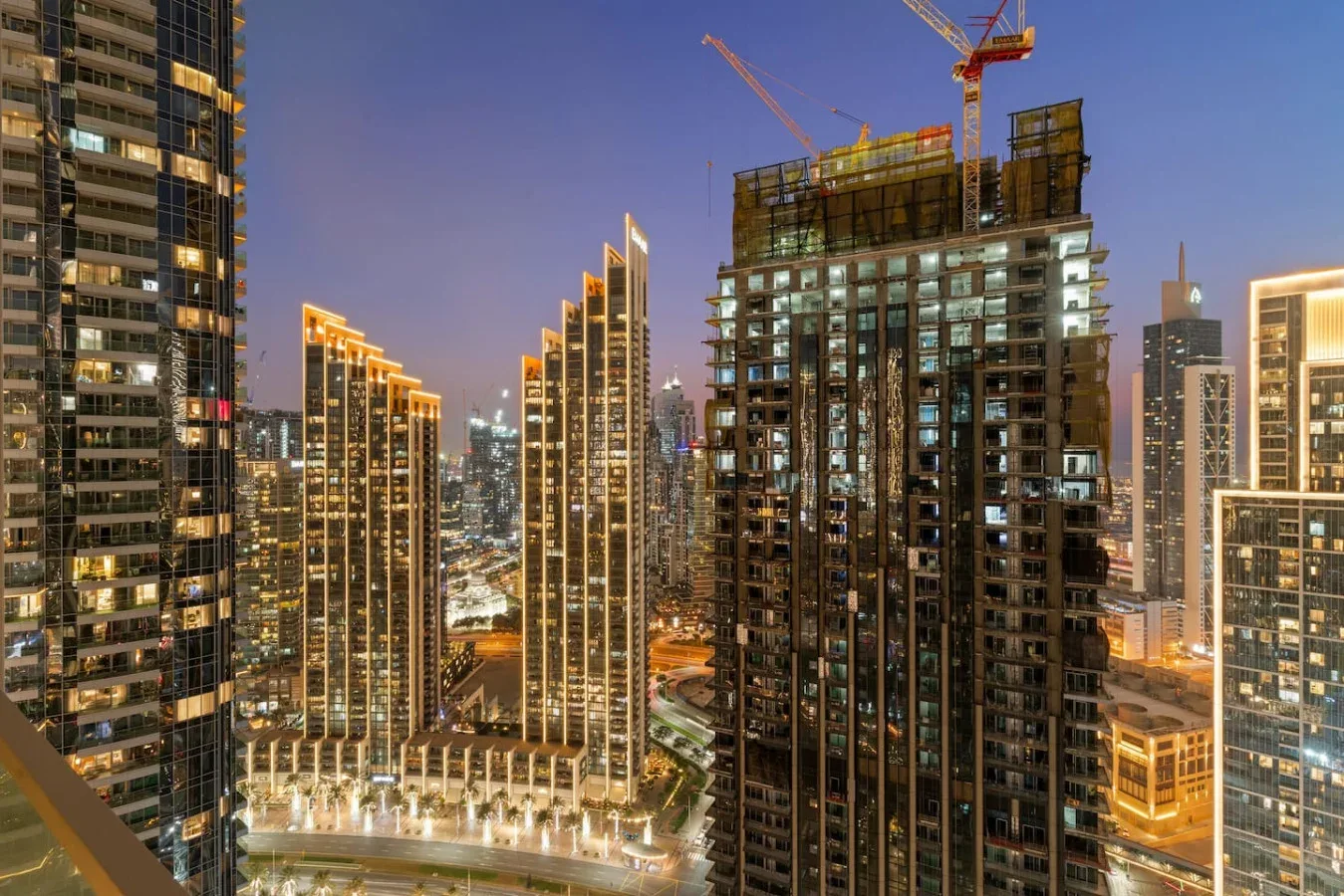 Un paisaje urbano nocturno muestra edificios altos iluminados y grúas de construcción.