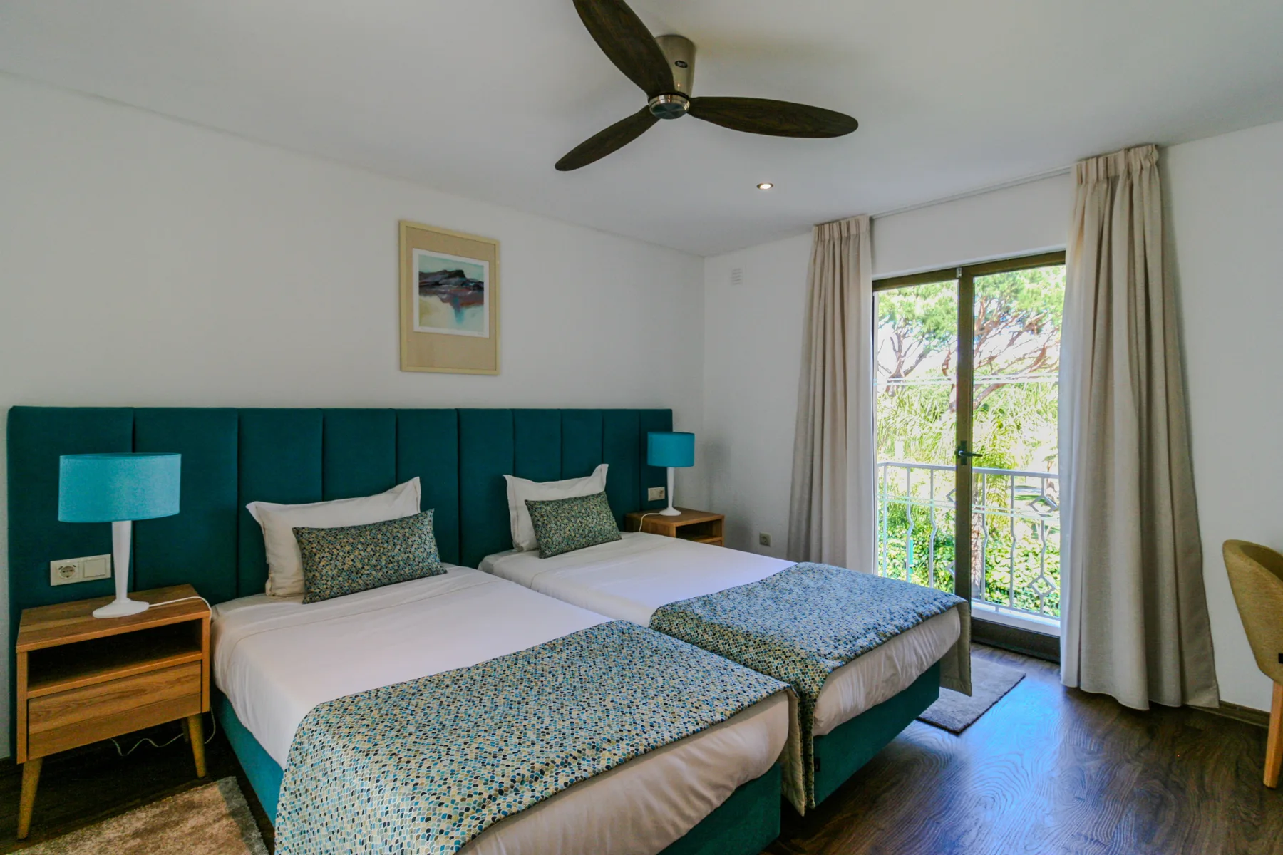 A bedroom with two beds, teal headboard, and a glass door opening to a balcony.