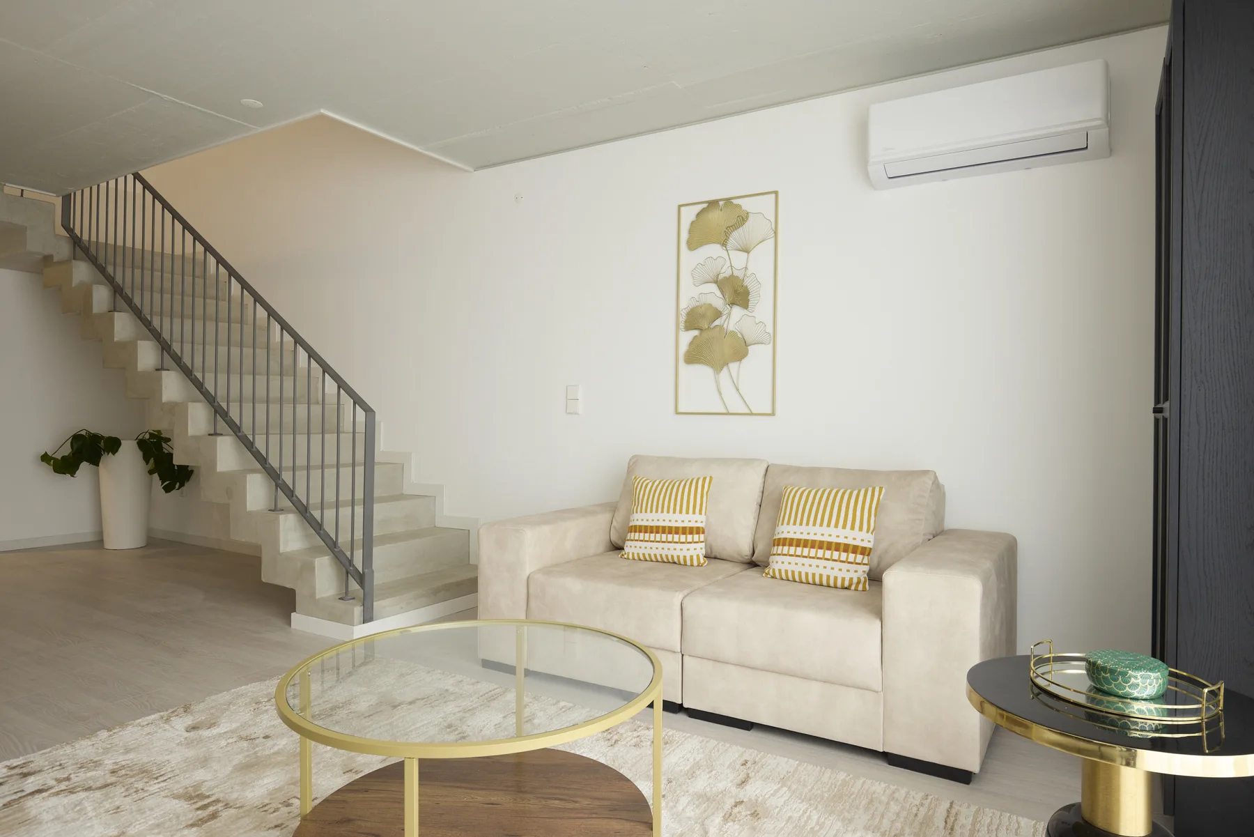 Una sala de estar con sofá beige, mesa de centro de vidrio y escalera.