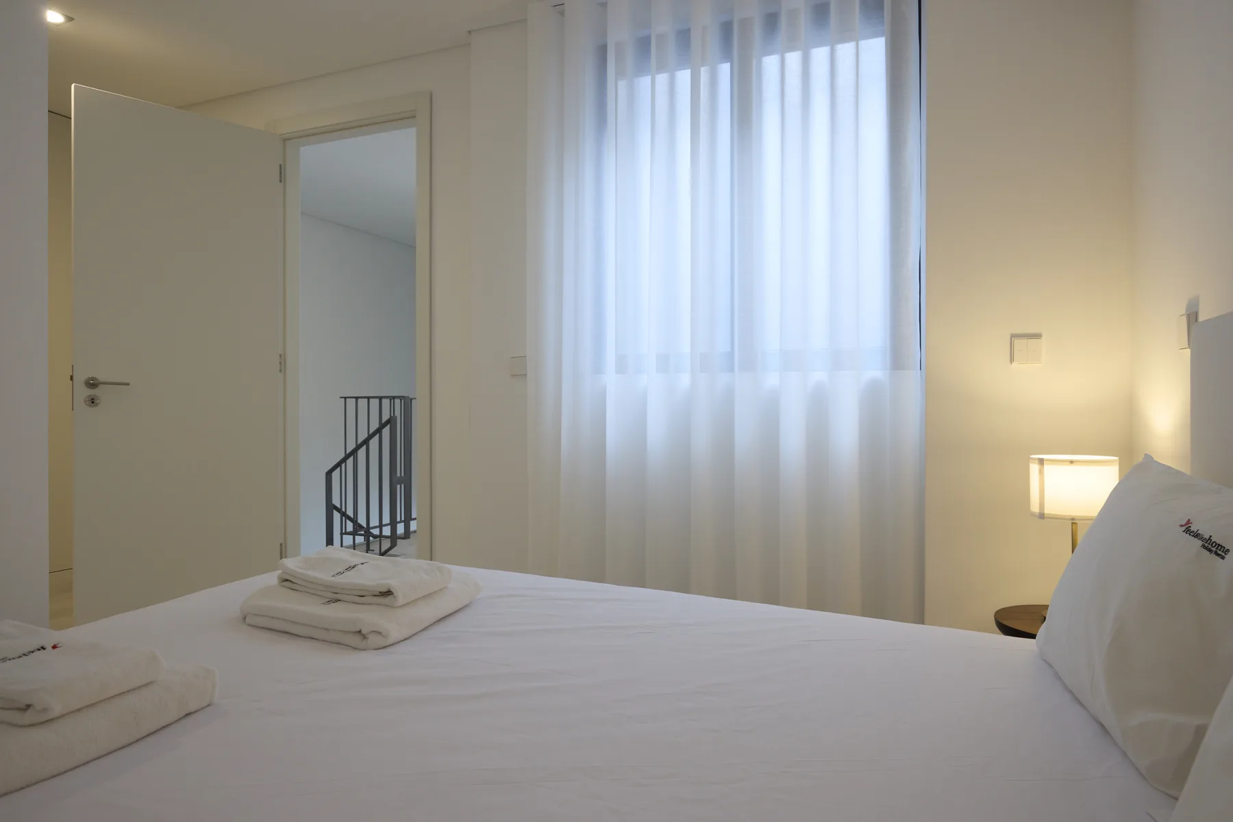 Un dormitorio con una cama hecha, toallas dobladas y una ventana con cortinas translúcidas.