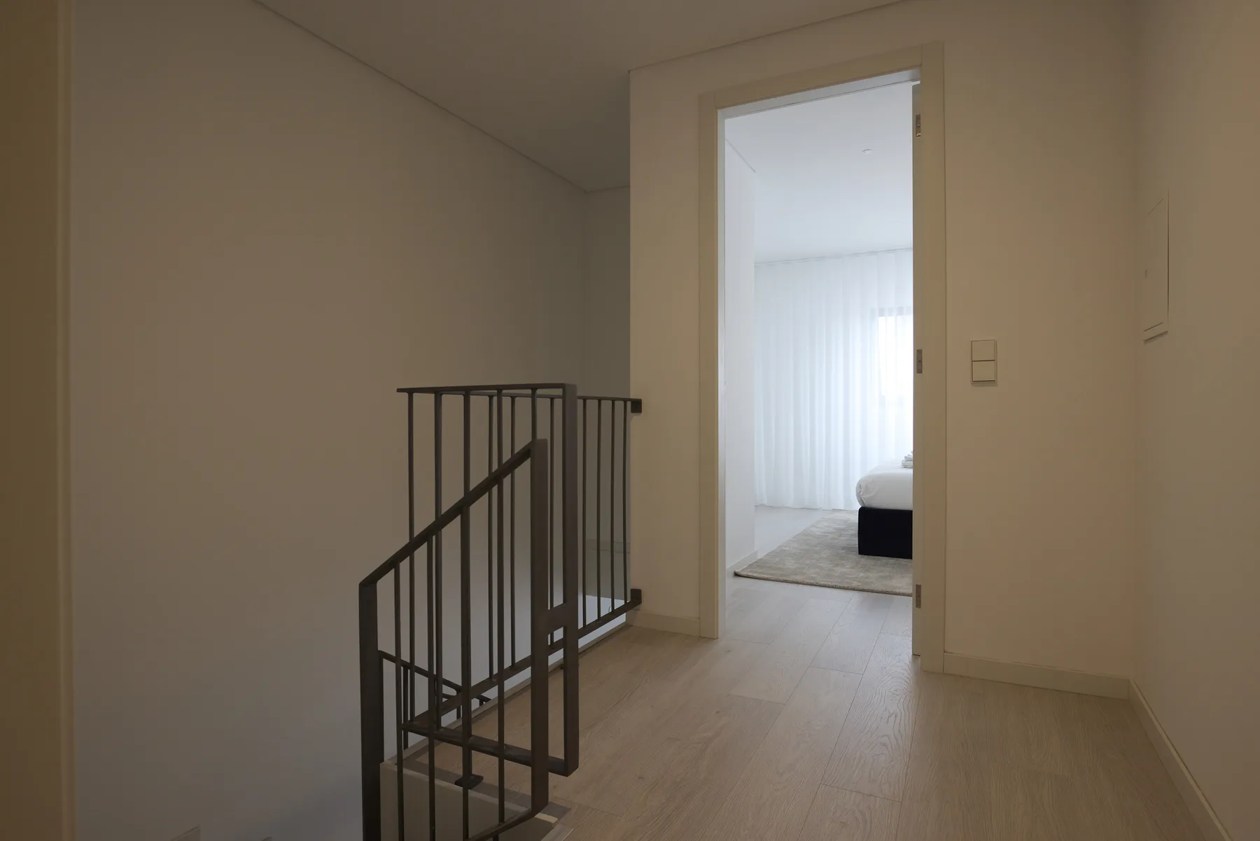 Un pasillo luminoso da a un dormitorio, con una barandilla oscura en primer plano.