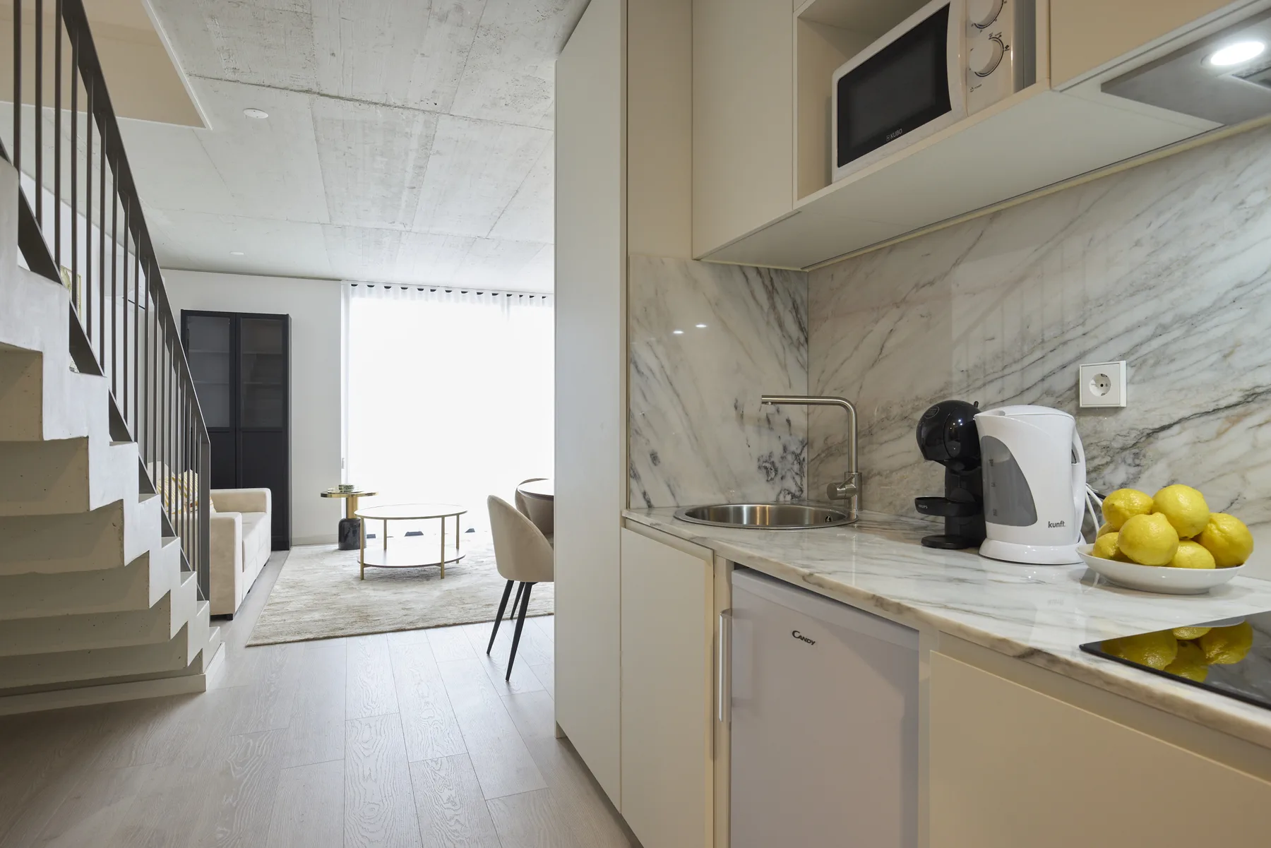 Una luminosa sala de estar de planta abierta con una escalera y una encimera de cocina compacta.