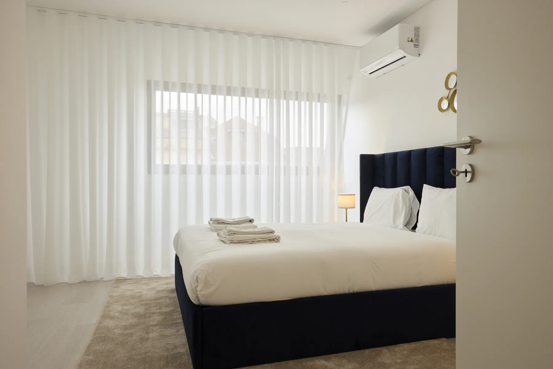 Un dormitorio luminoso con una cama bien hecha y cortinas translúcidas frente a una ventana amplia.