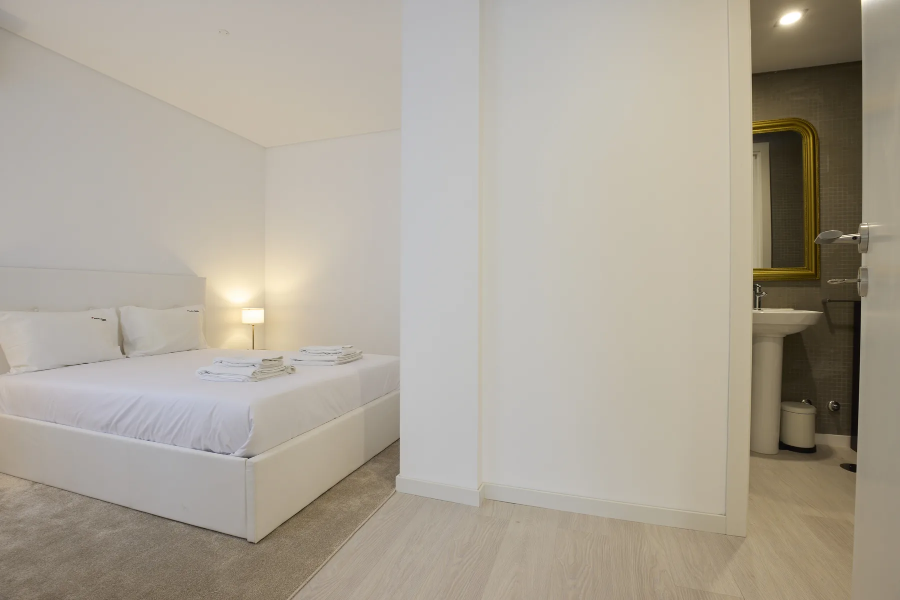 A bright bedroom with a white bed beside an open bathroom.