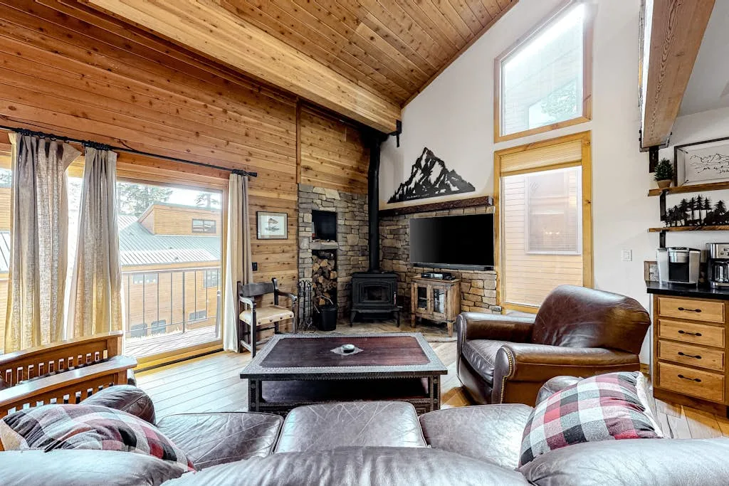 A wood-paneled living room with a television, stone fireplace, and large windows.
