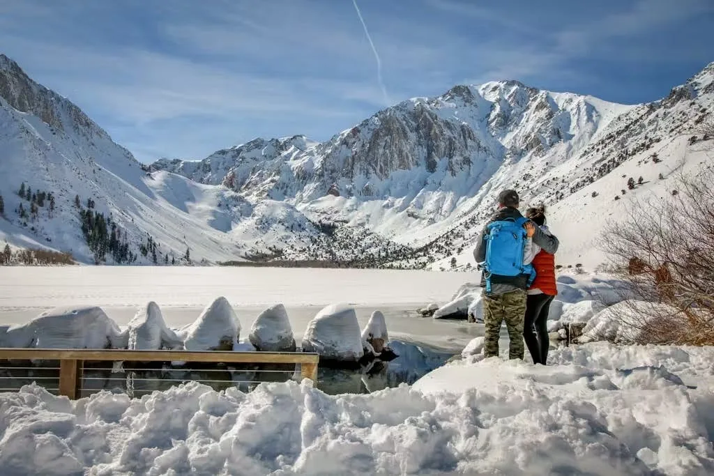 Two people stand beside a snowy lake beneath a mountain range.
