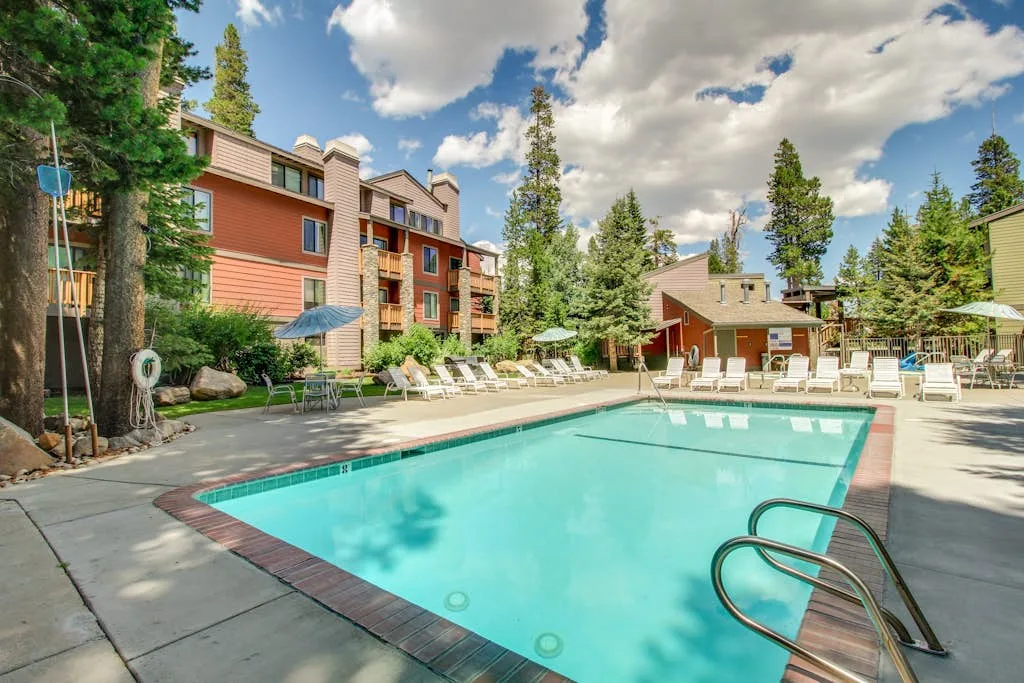 An outdoor swimming pool sits beside multi-story buildings and surrounding trees.