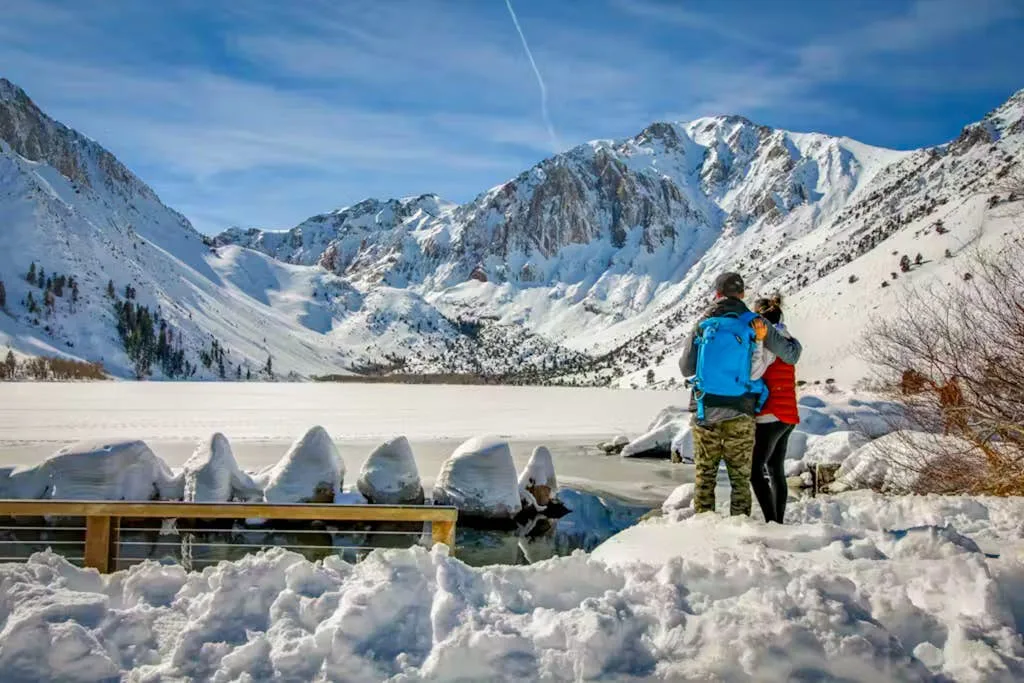 Two people stand beside a snow-covered mountain lake beneath rugged peaks.