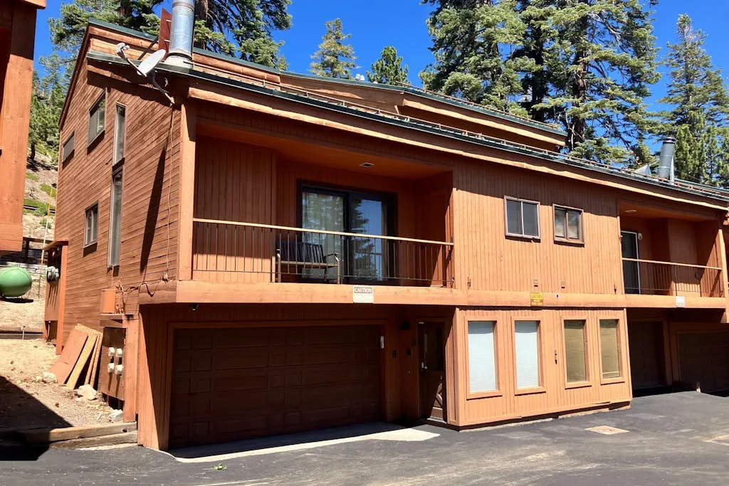 A wooden mountain home with a balcony above the garage, surrounded by tall trees.