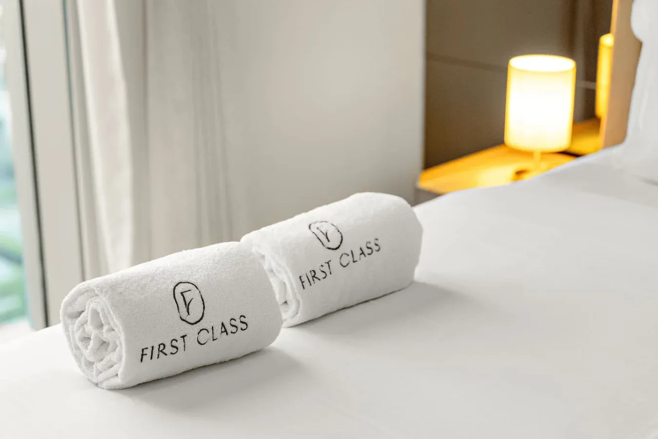 Una cama bien tendida con toallas enrolladas junto a una lámpara encendida.