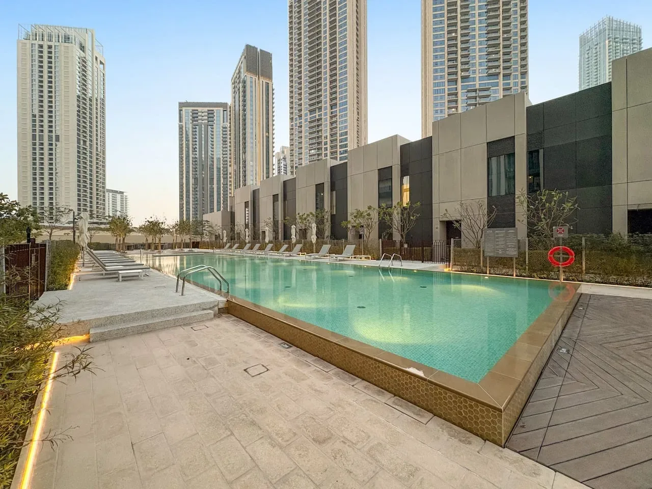 Una piscina al aire libre está junto a una terraza pavimentada, con edificios altos al fondo.