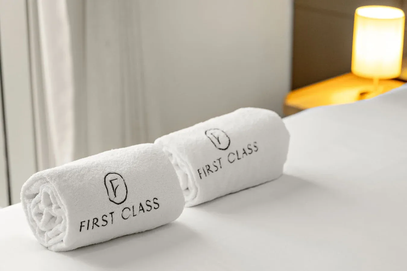 Toallas blancas enrolladas sobre una cama junto a una lámpara encendida.