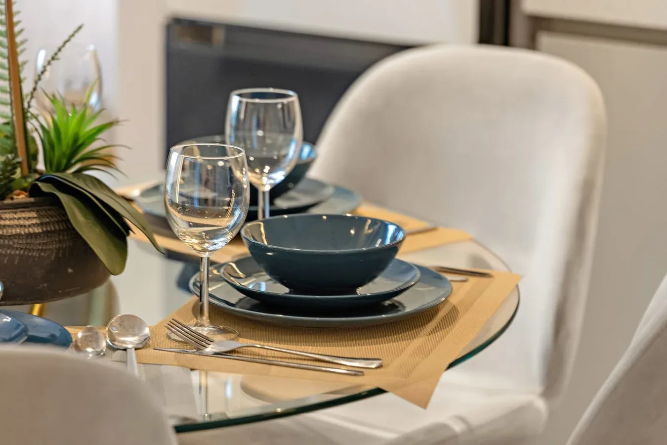 Una mesa de comedor está puesta con platos, cristalería y un cuenco junto a una planta.