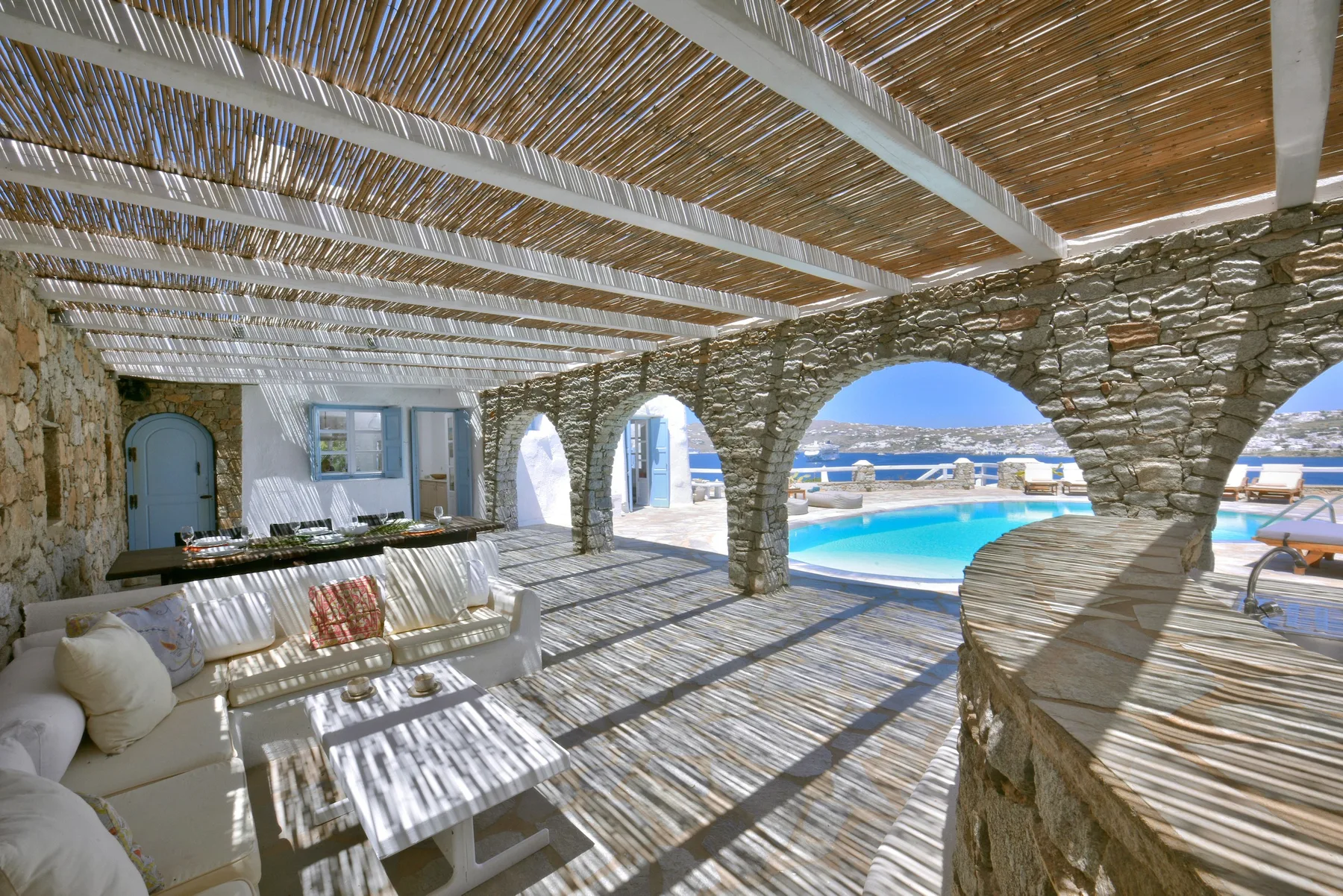 A shaded stone terrace with seating overlooks a swimming pool and the sea.
