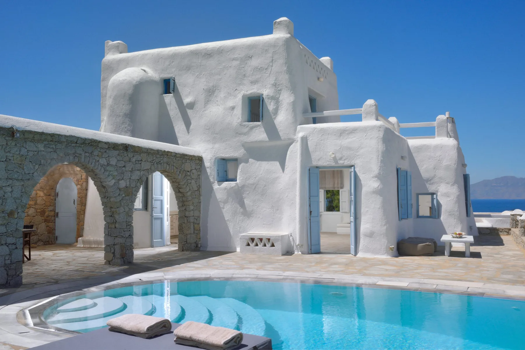 A whitewashed villa stands beside a turquoise swimming pool under a clear blue sky.