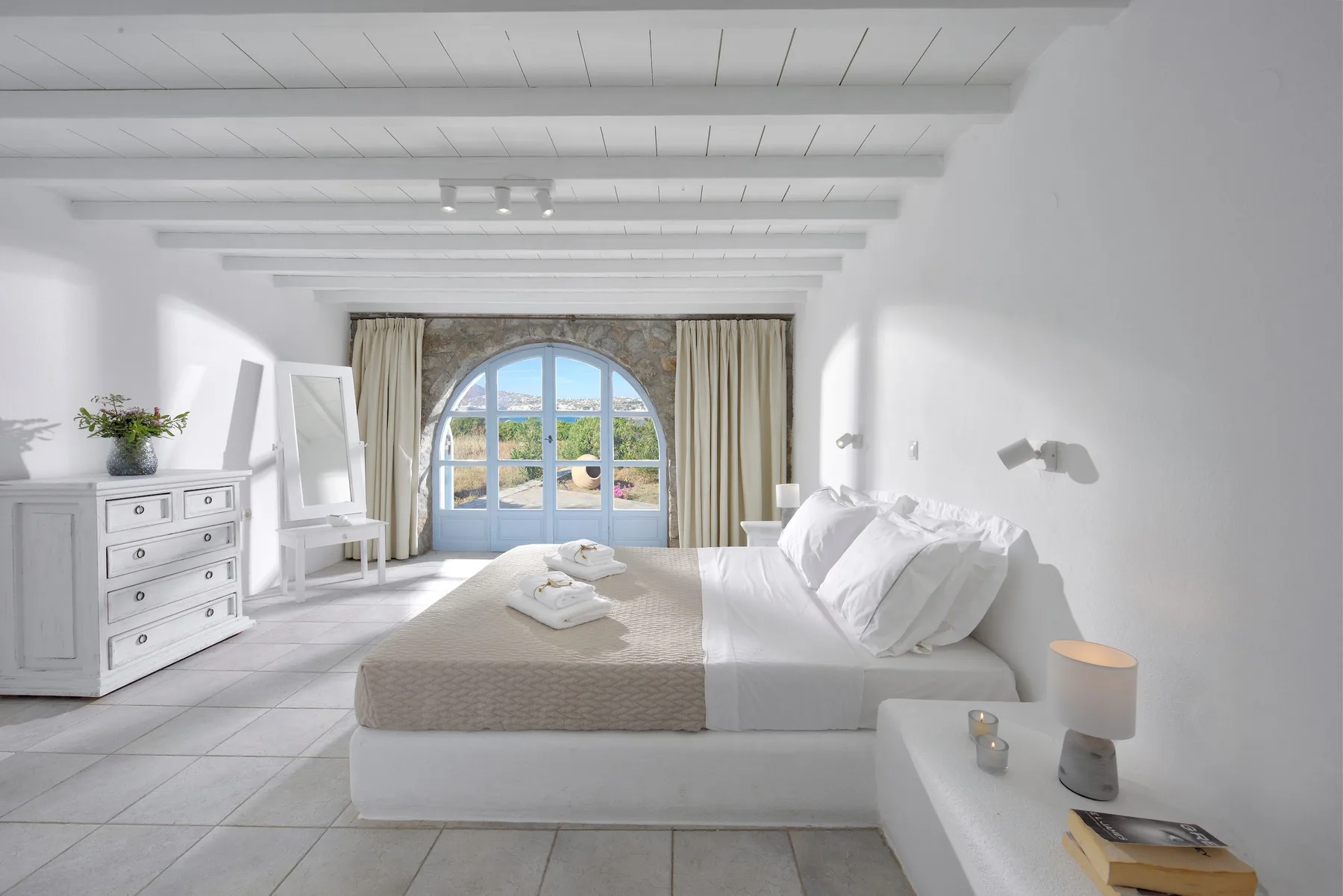 Un dormitorio luminoso tiene una cama grande bajo vigas blancas y una ventana arqueada.